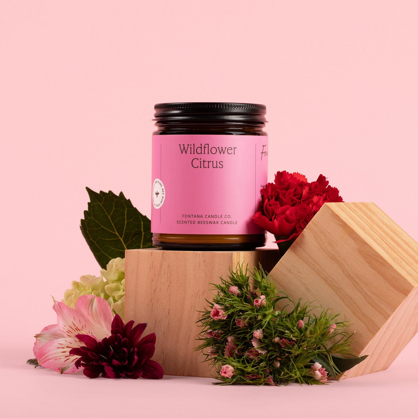 Amber glass candle jar labeled 'Wildflower Citrus' with flowers and leaves on a pink background