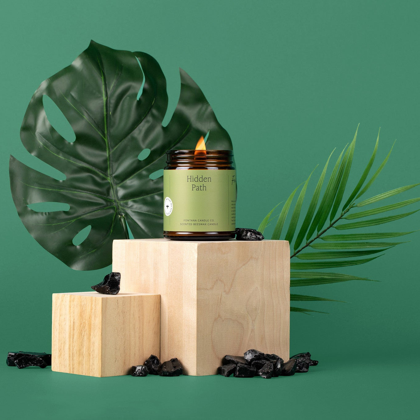 Lit amber glass jar candle with a green label on a wooden block with palm leaves against a green background.
