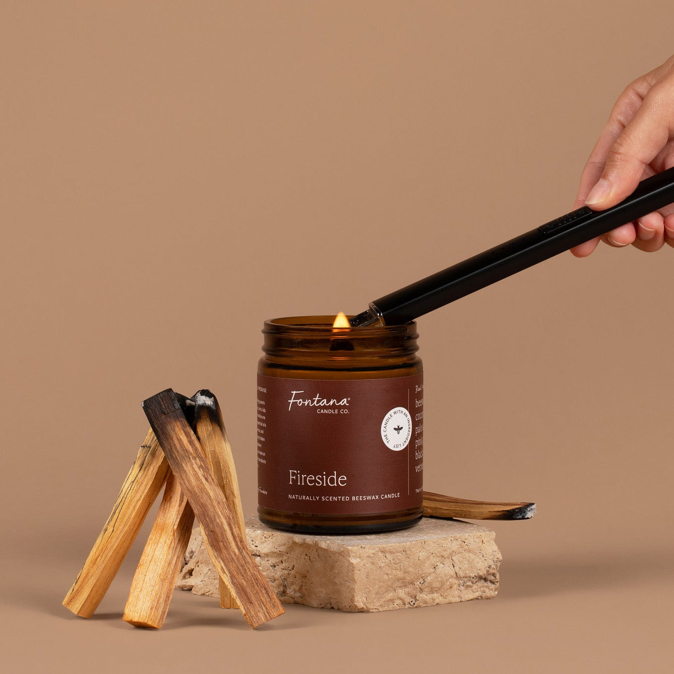 An amber glass jar candle labeled 'Fireside', lit with a black lighter held by a hand, styled with palo santo wood sticks on a stone base against a solid tan background.