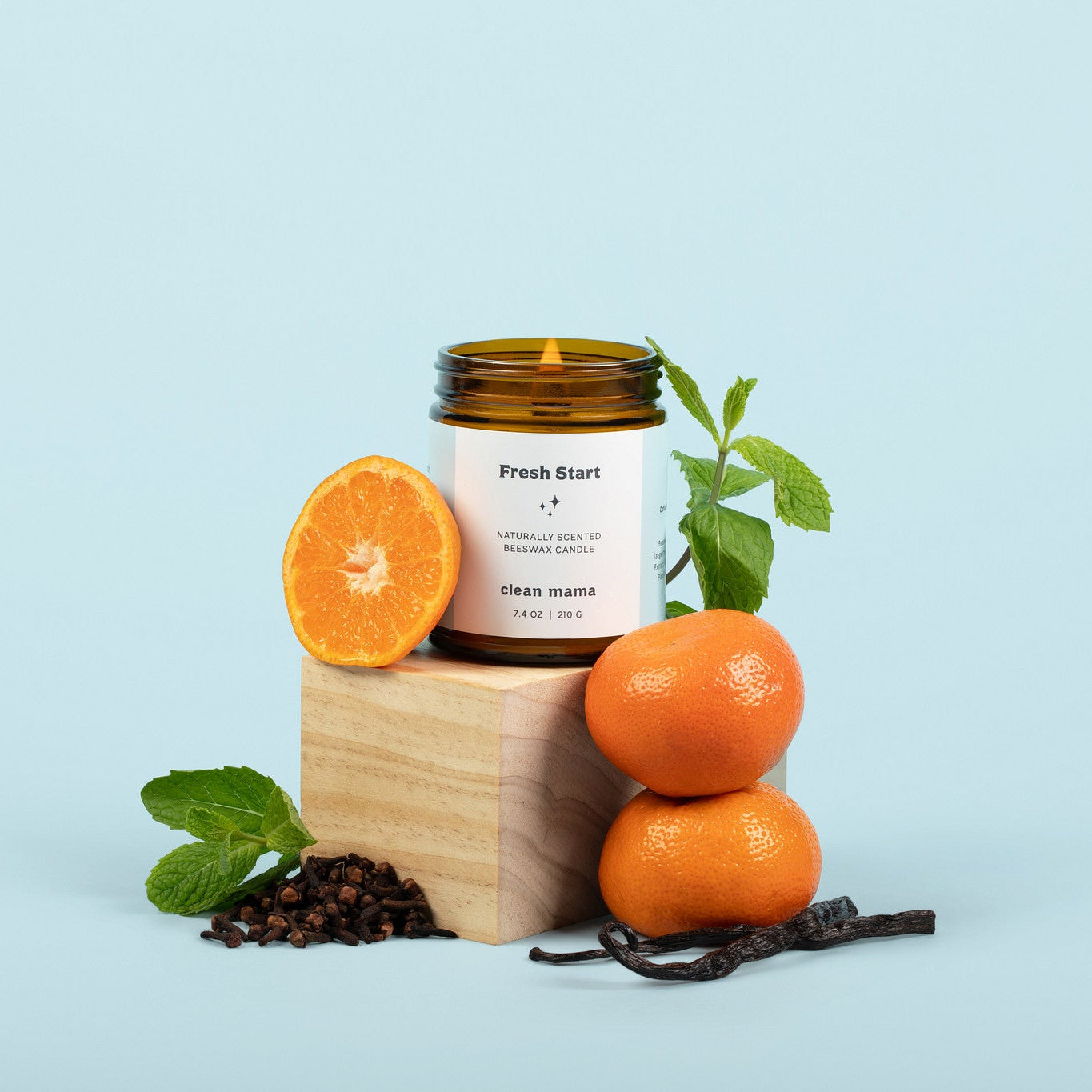 Lit amber glass 'Fresh Start' candle in front of a light blue background, surrounded by oranges, mint, cloves, and vanilla.