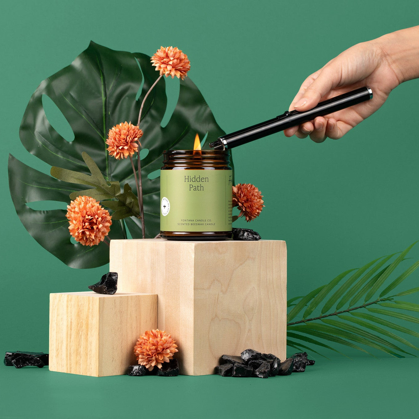 Hand lighting an amber glass jar candle with a green label on a wooden block with green leaves and flowers in the background.