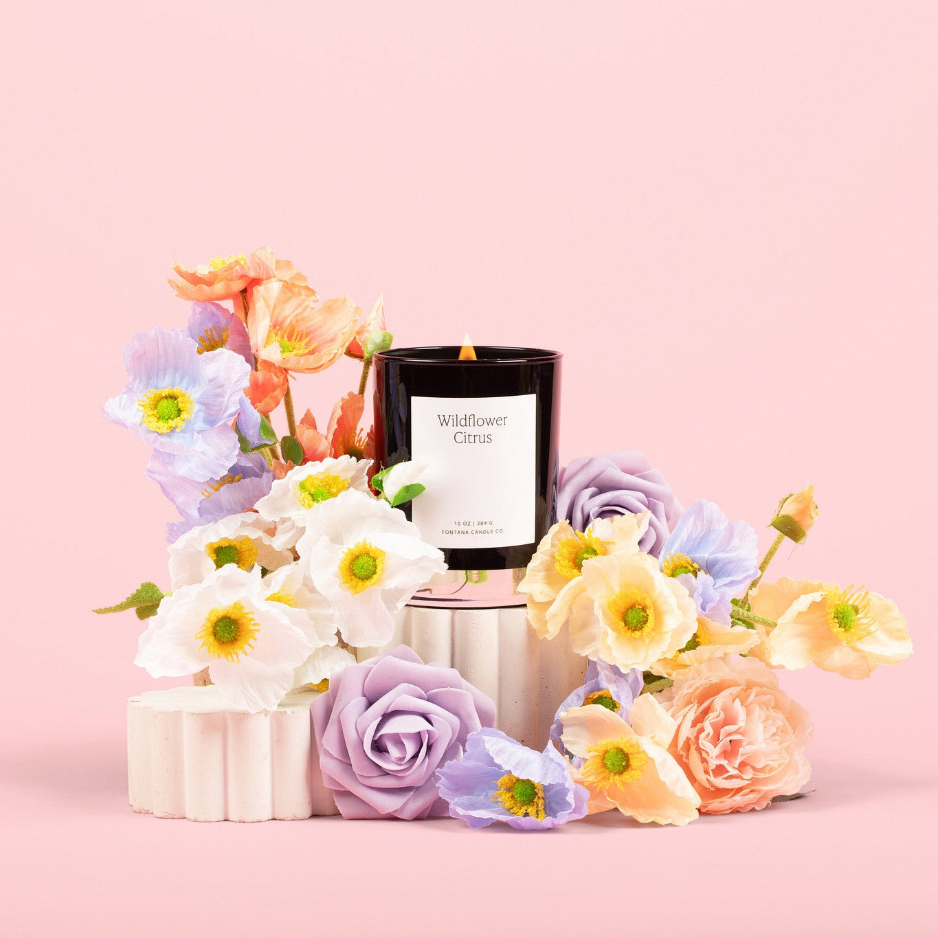 Lit black glass candle in front of a pink background, surrounded by pastel colored flowers.