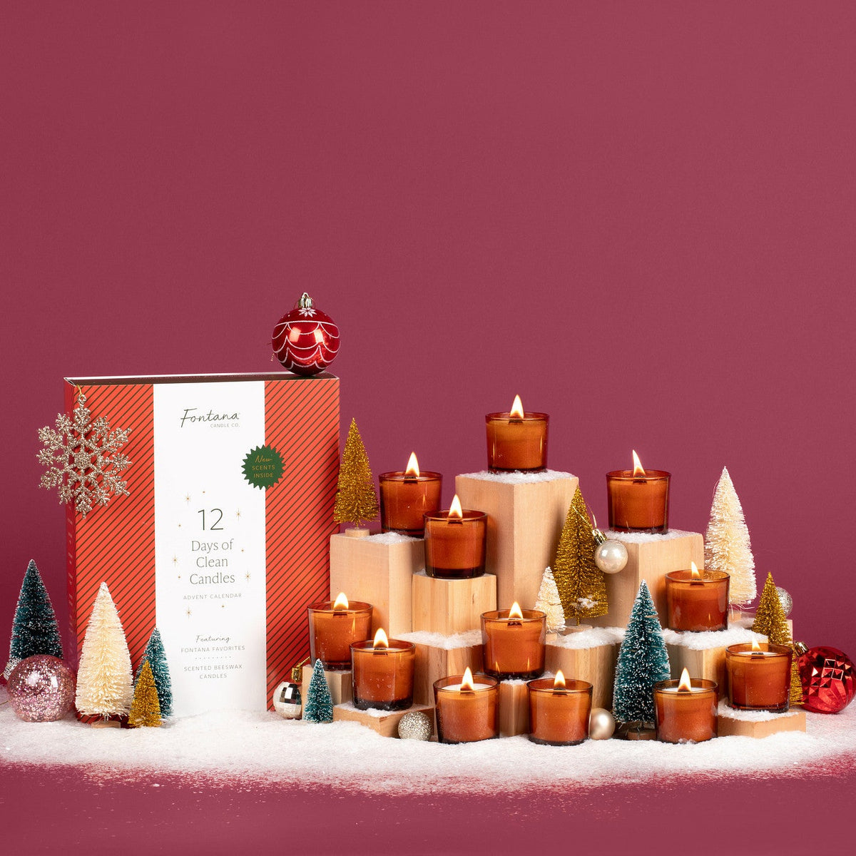 12 Lit amber glass mini candles in front of a festive plum colored background, surrounded by fake snow, holiday ornaments, mini Christmas trees, and the 12 Days of Clean Candles box. 