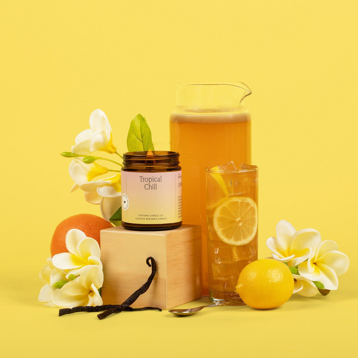 Lit amber glass jar candle with a bright label in front of a yellow background with a pitcher and glass of electrolytes. It is surrounded by white flowers and lemons, and vanilla pods. 