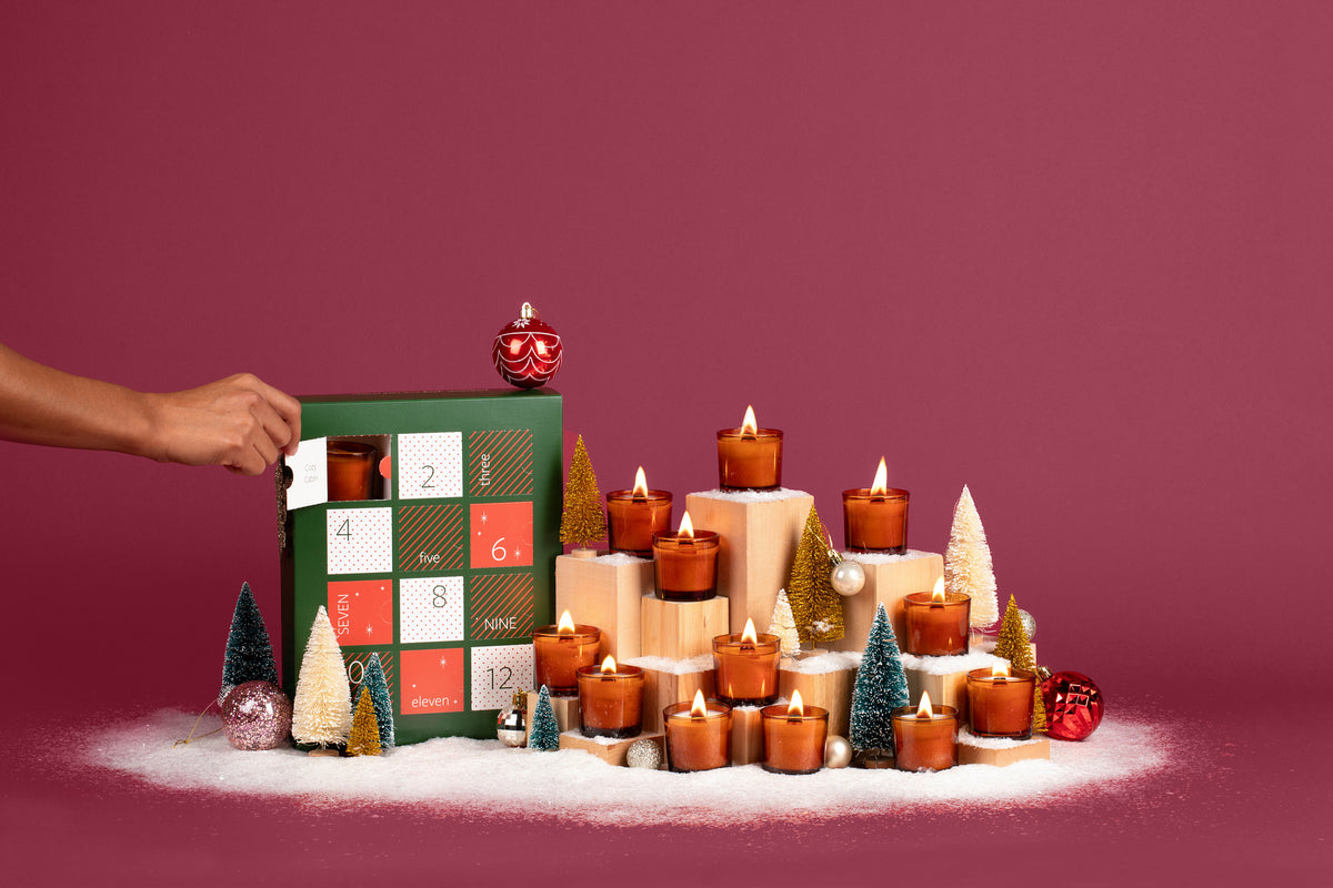 12 Lit amber glass mini candles in front of a festive plum colored background, surrounded by fake snow, holiday ornaments, mini Christmas trees, and the 12 Days of Clean Candles box. 