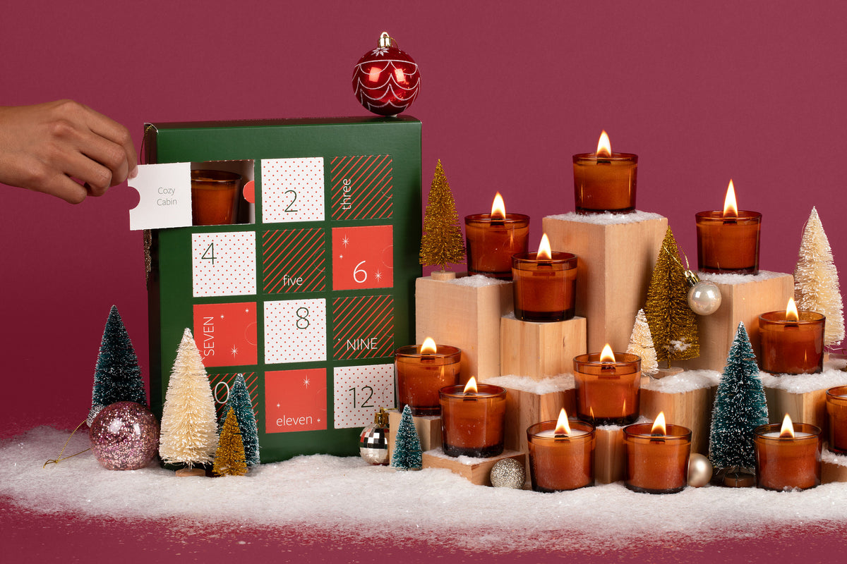 12 Lit amber glass mini candles in front of a festive plum colored background, surrounded by fake snow, holiday ornaments, mini Christmas trees, and the 12 Days of Clean Candles box. 