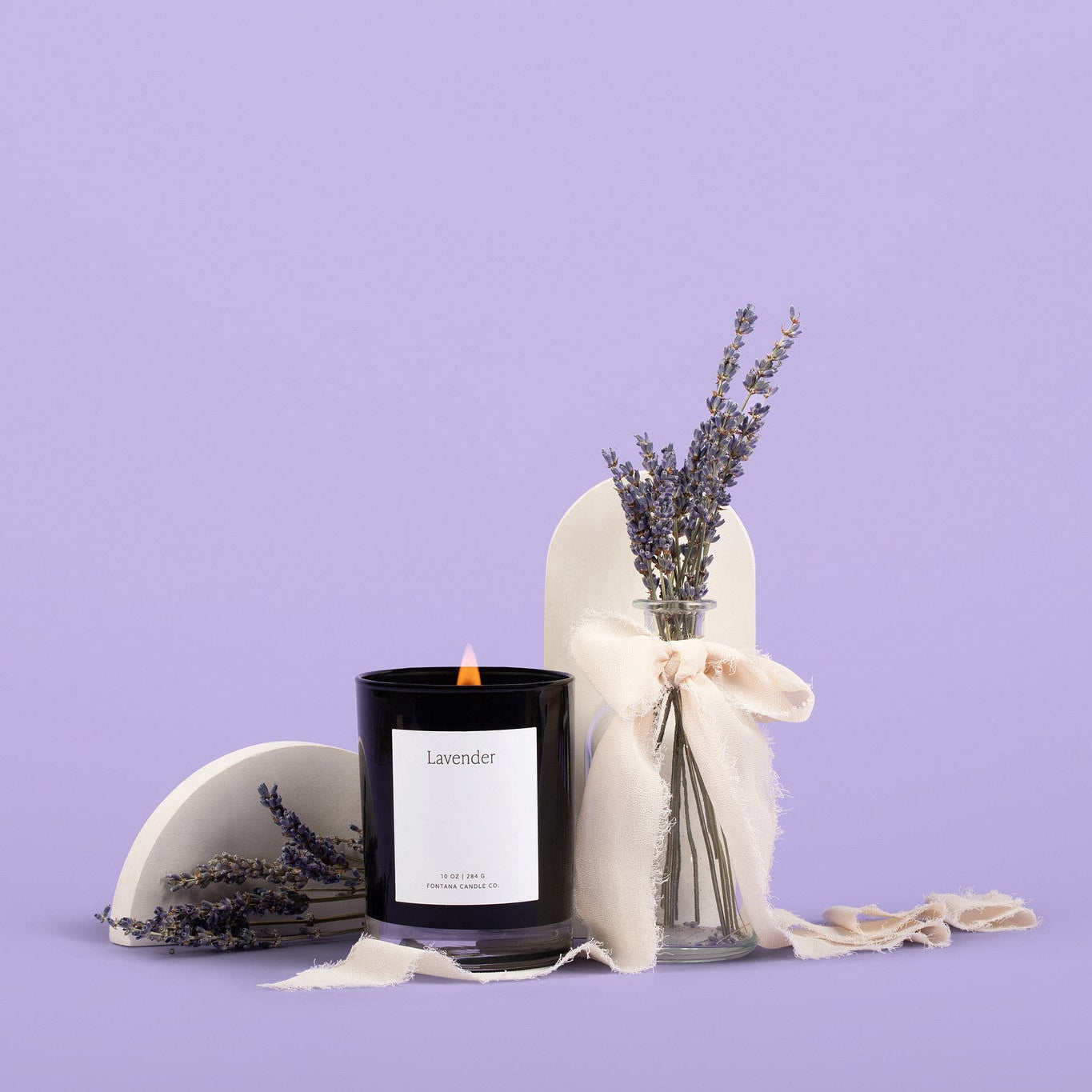 Lit black glass candle with a white label on a purple background, surrounded by lavender flowers.