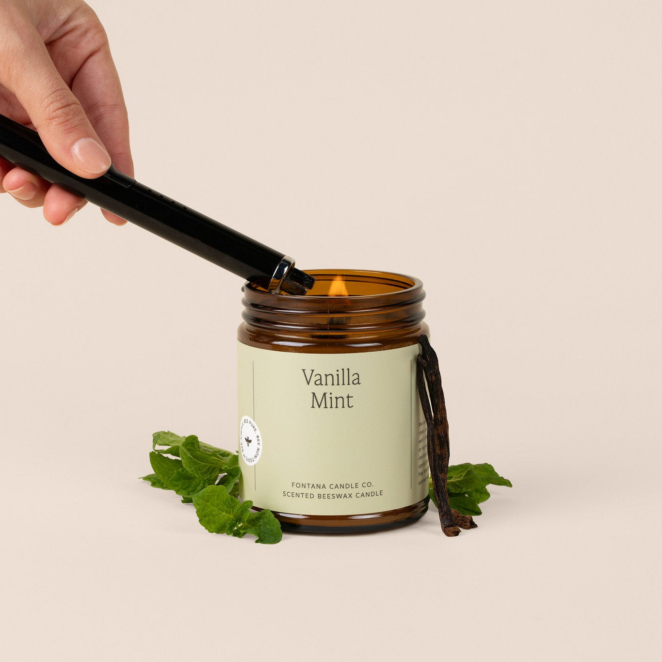 Hand lighting an amber glass jar candle labeled 'Vanilla Mint' with a beige background.