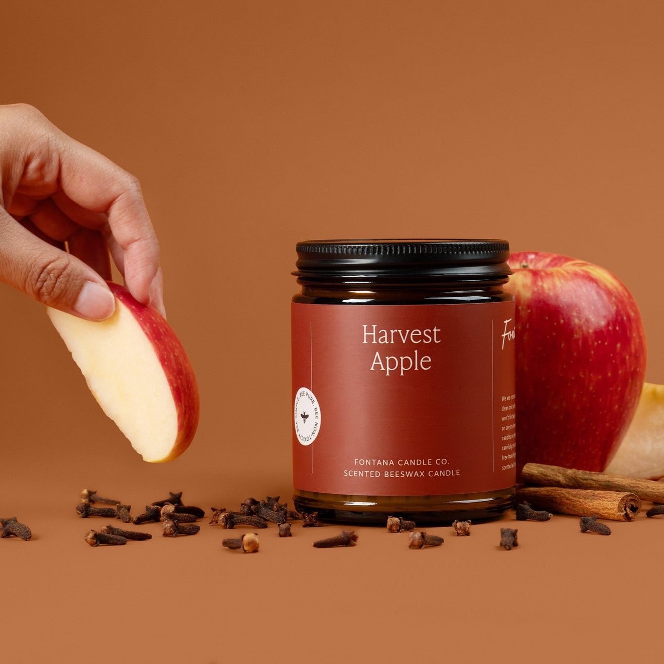 Amber glass jar candle of 'Harvest Apple' candle with apple slices and cinnamon sticks on a brown background.