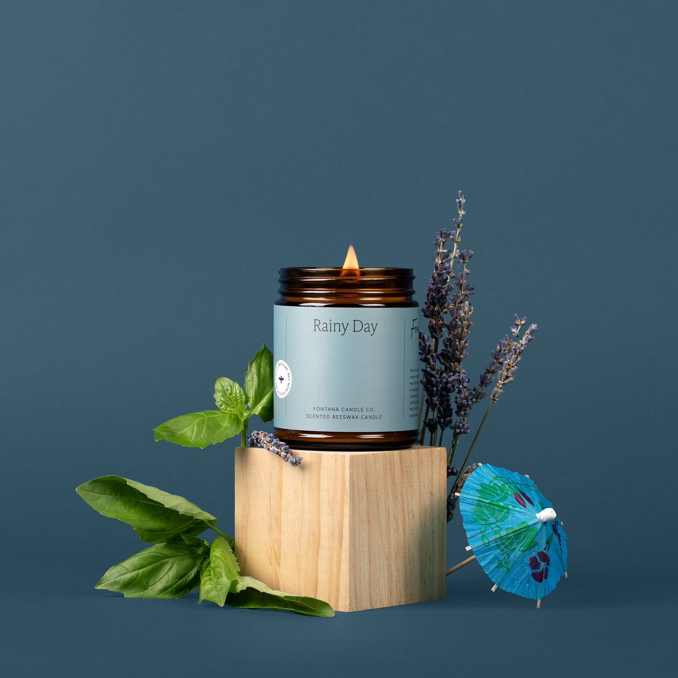 Lit amber glass jar candle with a blue label, in front of a blue background, surrounded by mint and lavender,  with a little blue drink umbrella.