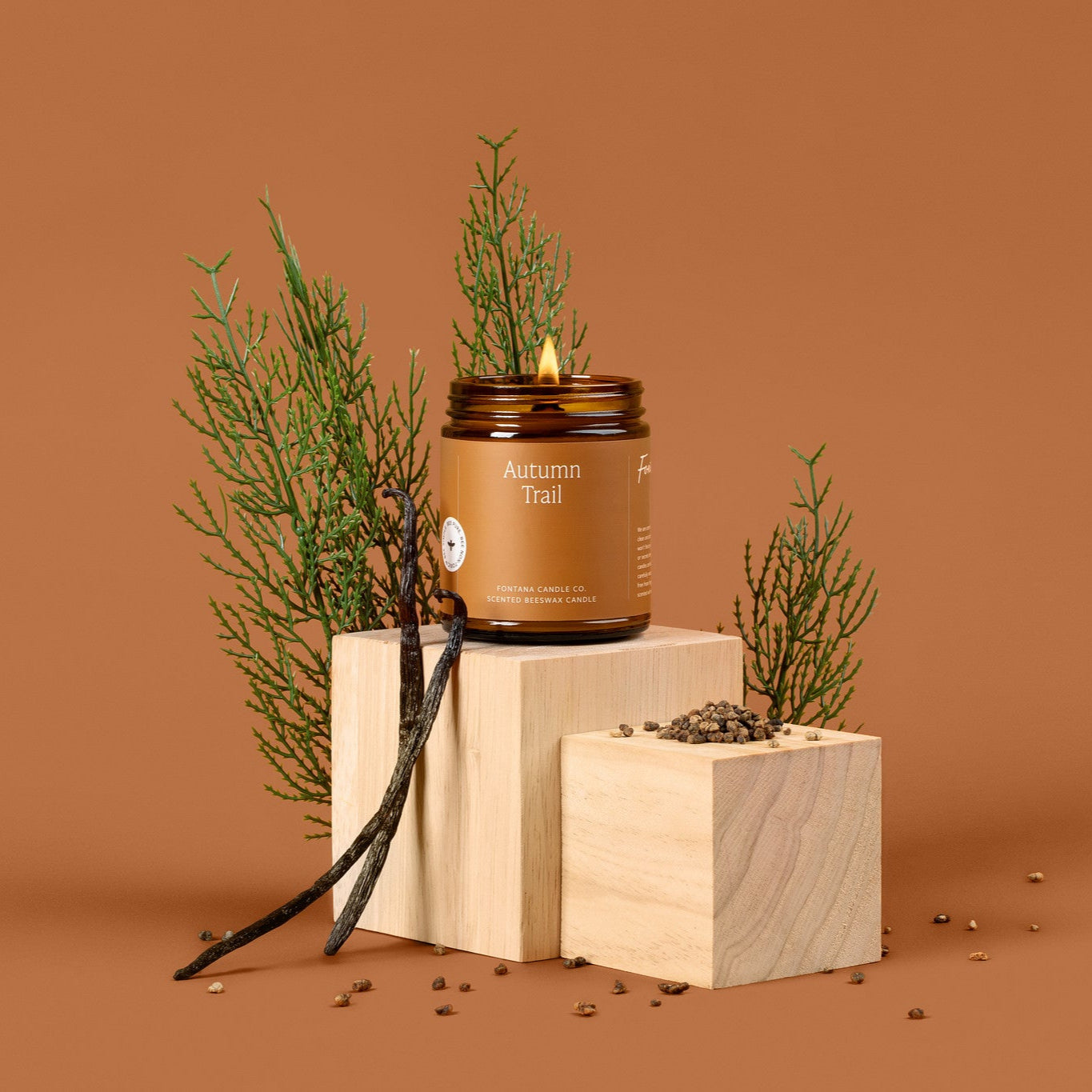 Lit amber glass jar candle labeled 'Autumn Forest' on a wooden block with greenery against a brown background.
