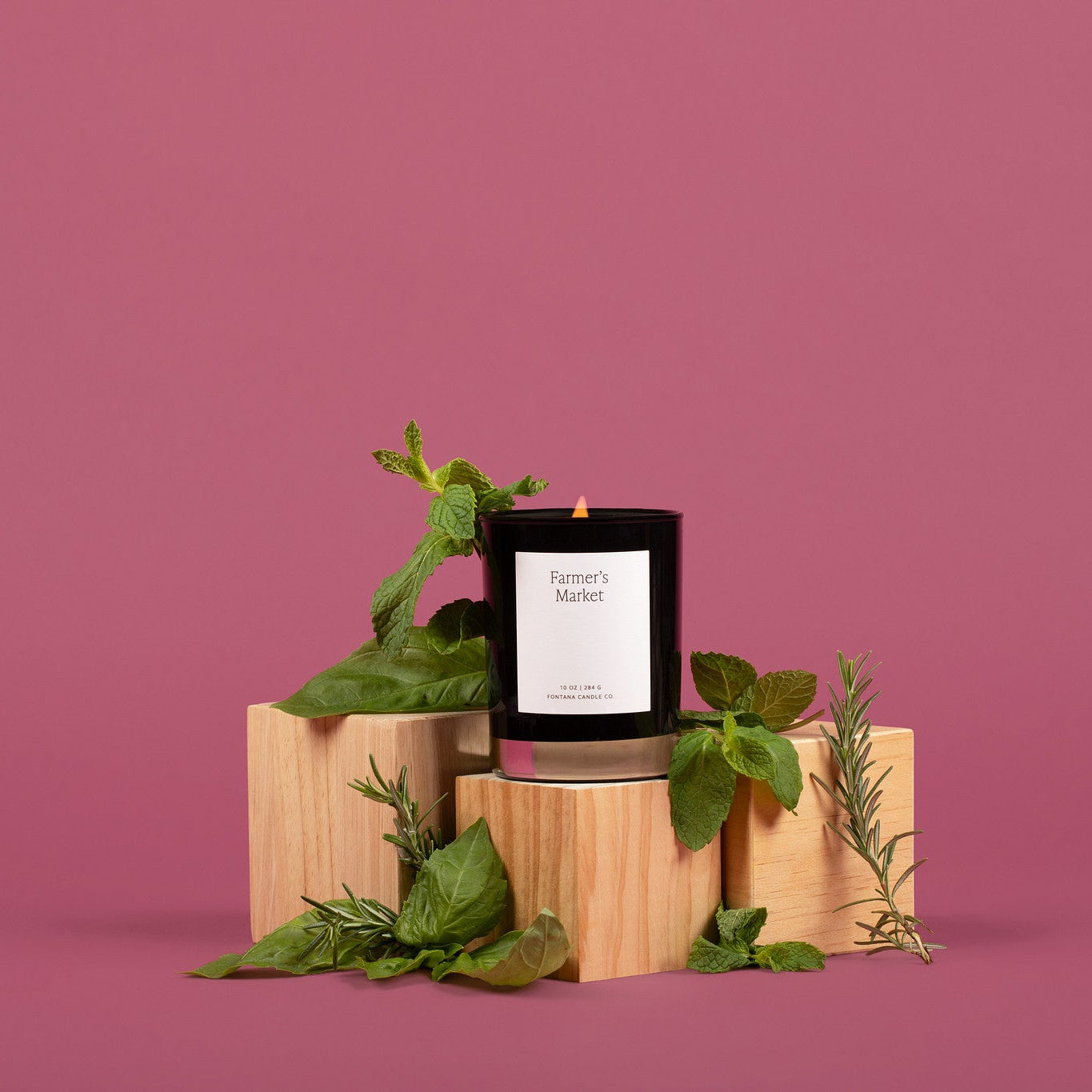 Lit glass candle with a white label on a wooden block, surrounded by basil, rosemary, and mint.