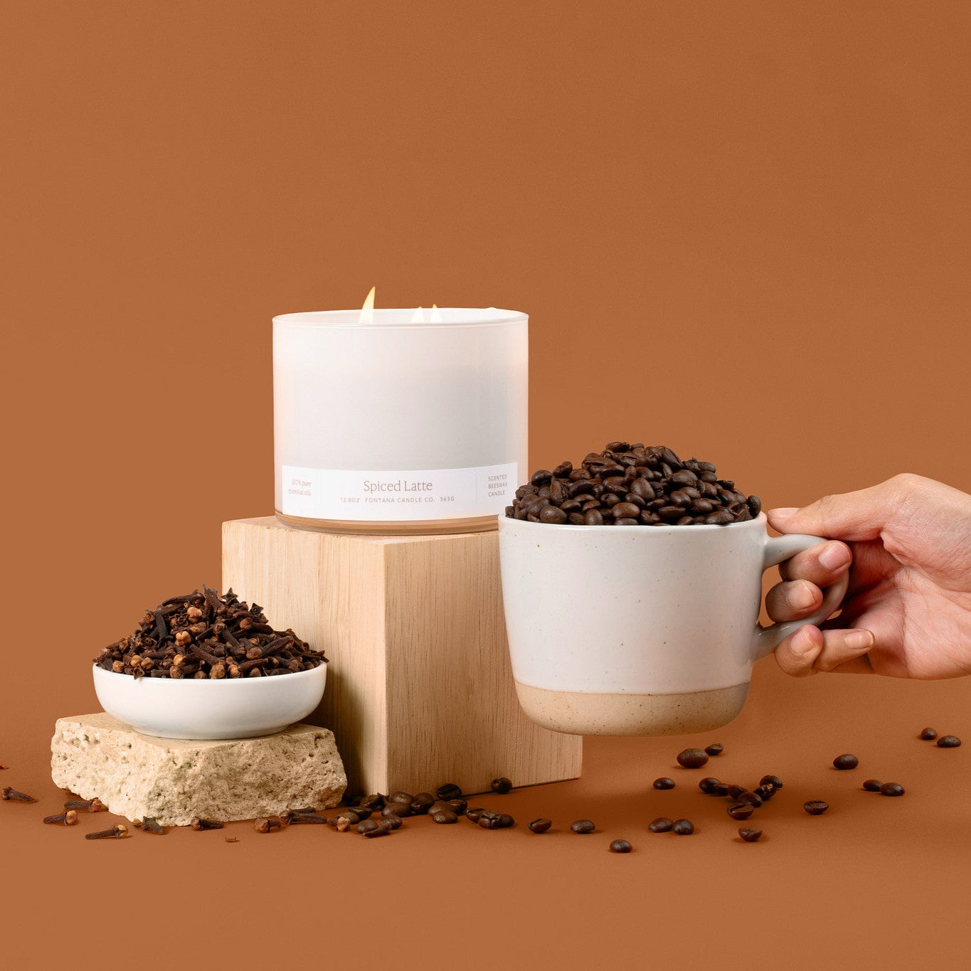 Lit 3-wick candle in a white glass vessel, surrounded by coffee beans, and a cup on a brown background.