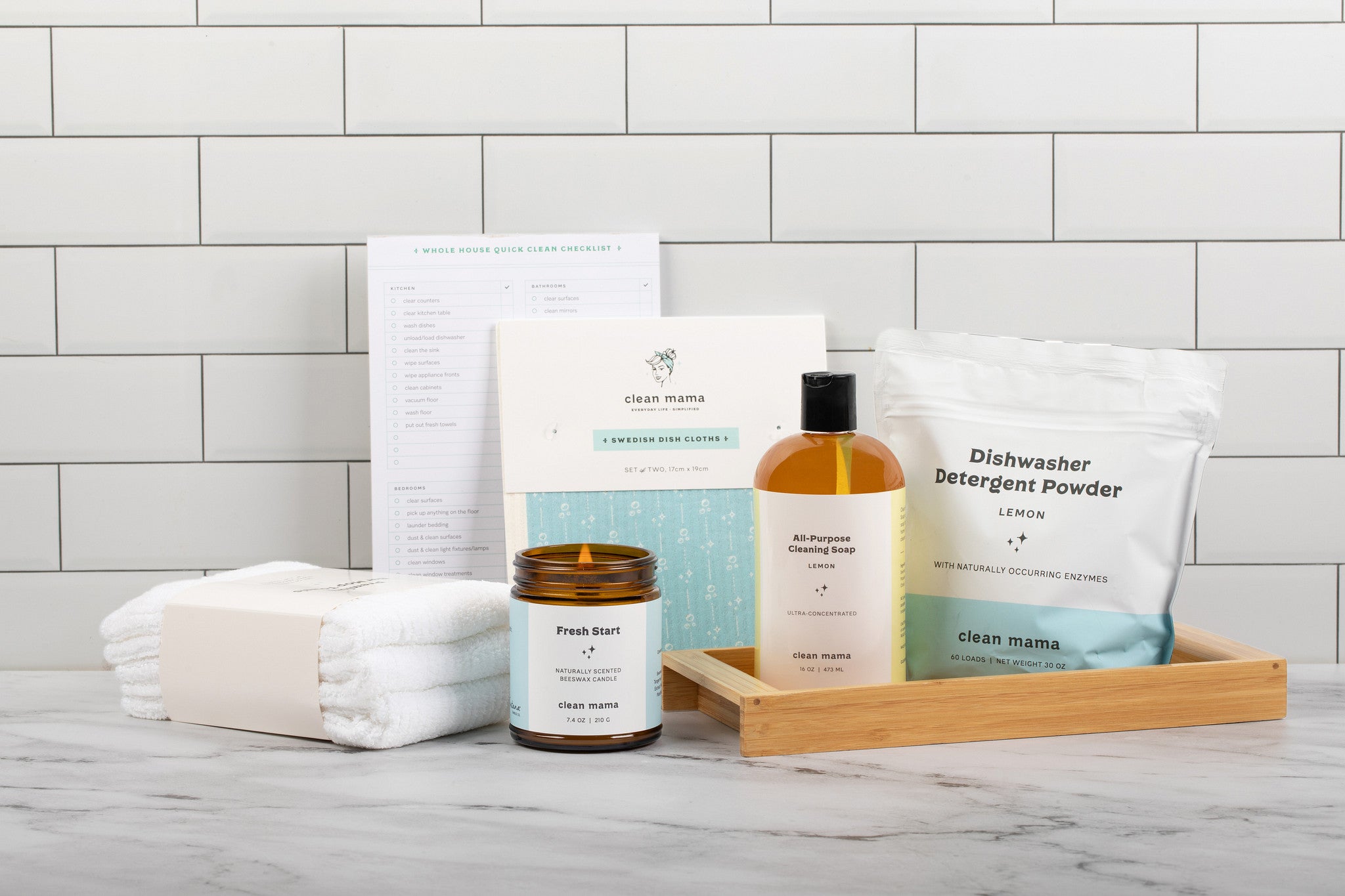 Lit amber glass jar candle with towels, all purpose cleaning soap, and dishwashing detergent power on a bathroom sink.