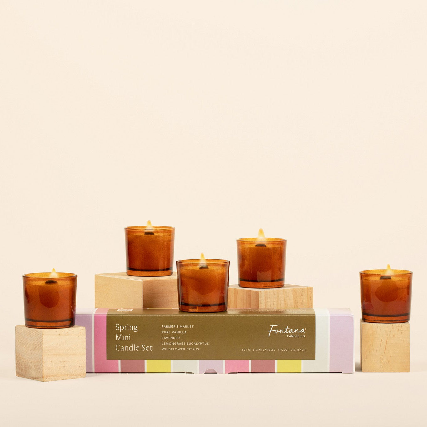 Set of 5 lit amber glass mini candles on wooden blocks in front of a cream background with the colorful spring gift box. 