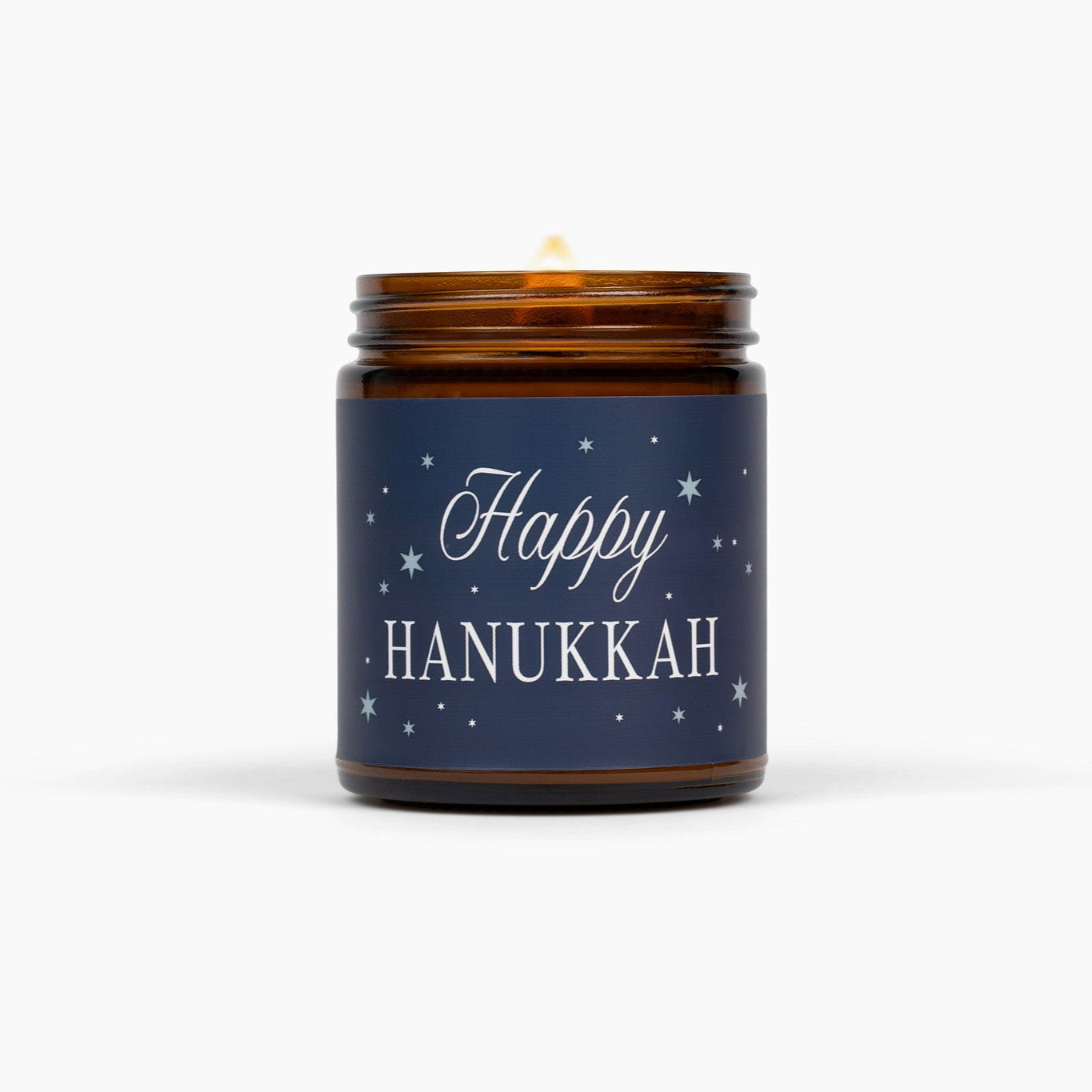 Lit amber glass jar candle with a dark blue label with Happy Hanukkah, in front of a white background.