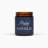 Lit amber glass jar candle with a dark blue label with Happy Hanukkah, in front of a white background.