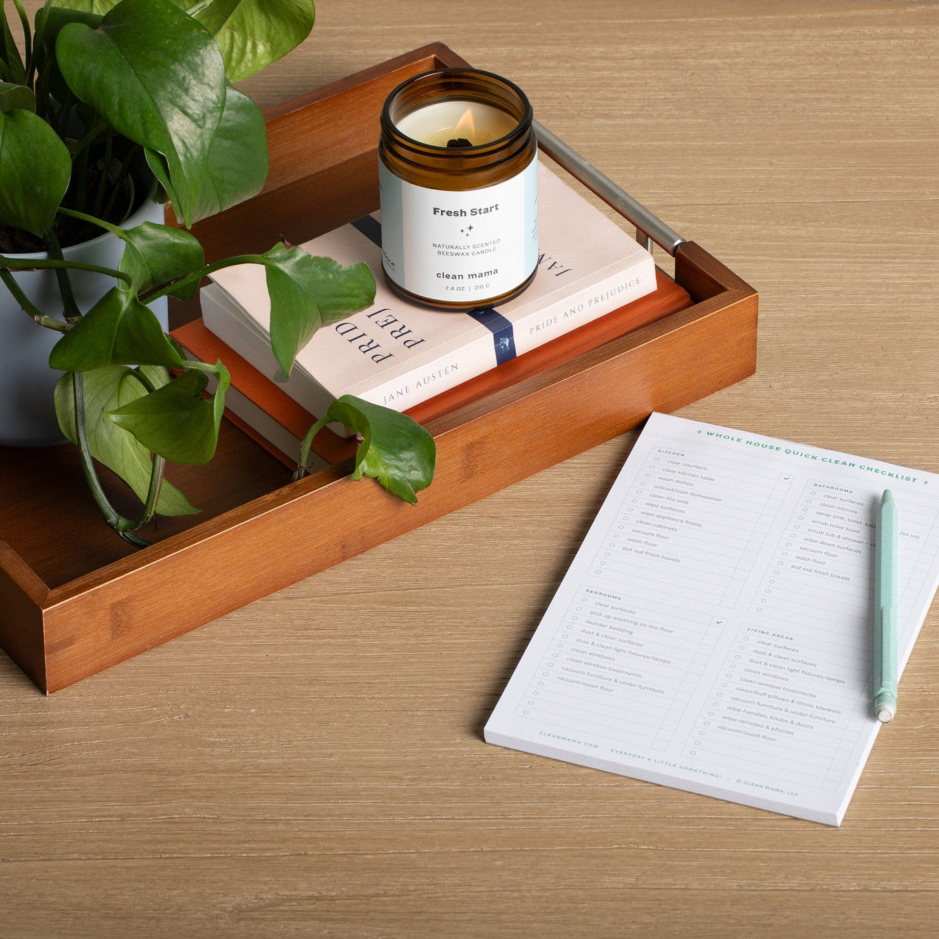 Wooden tray with a plant, lit 'Fresh Start' candle, and book on a wooden surface with a notepad and pen.