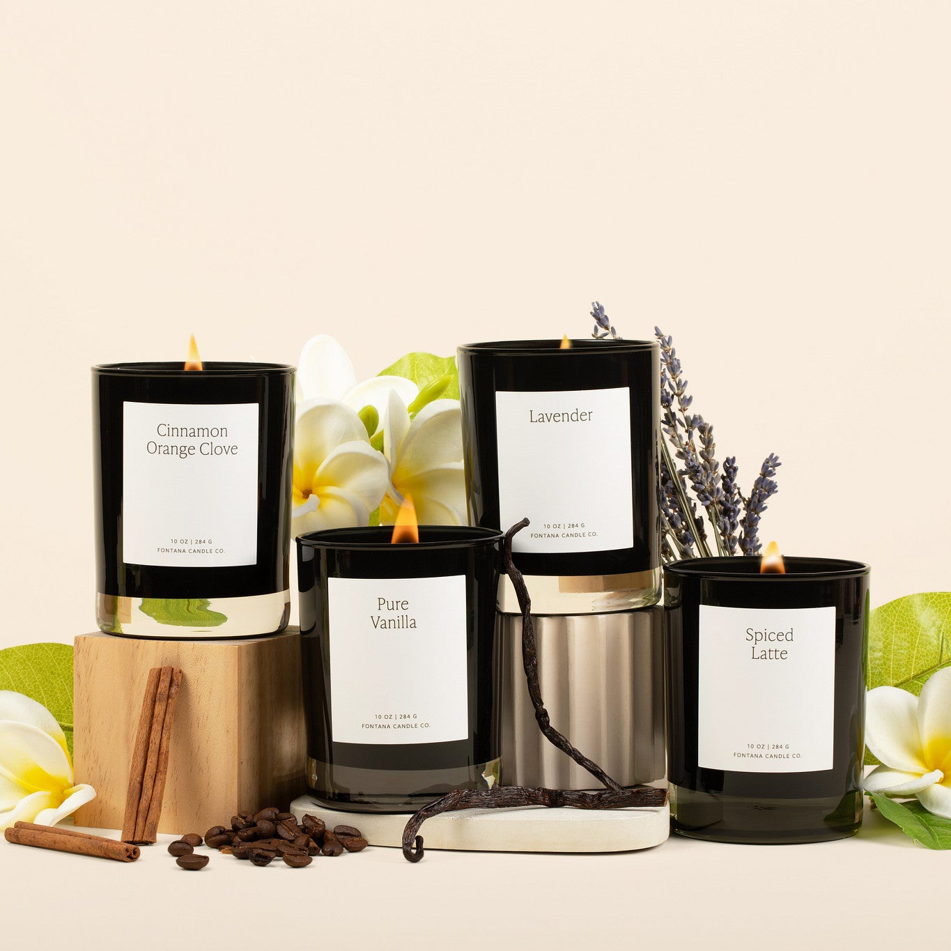 Four lit black glass candles on a cream background surrounded by flowers, spices, and vanilla. 