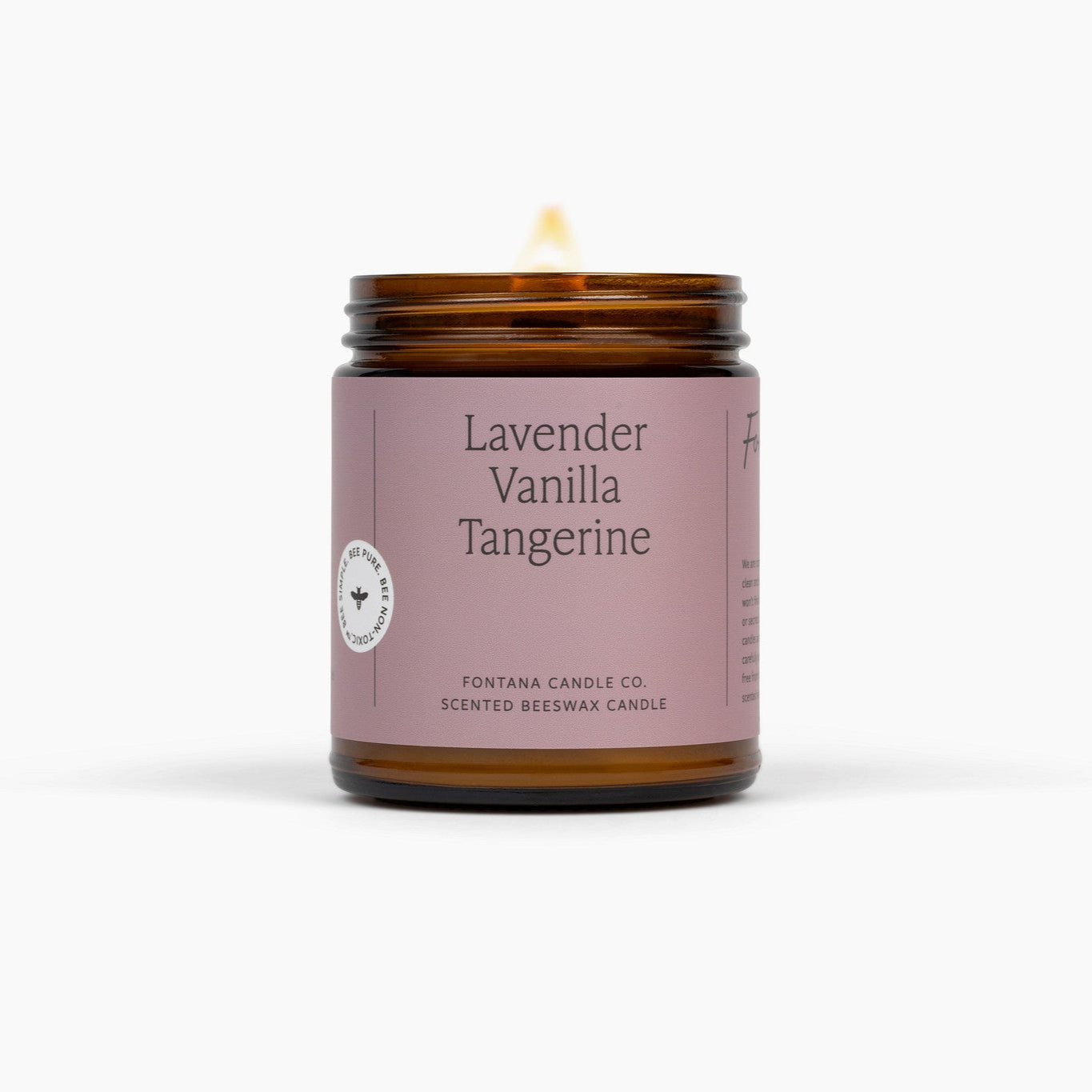 Lit amber glass jar candle with a light pink label that says Lavender Vanilla Tangerine. The candle is in front of a white background. 