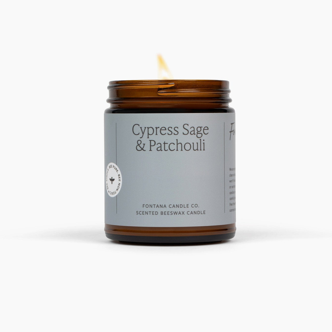 Lit amber glass jar candle with a light blue label called Cypress Sage & Patchouli, in front of a white background.