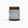 Lit amber glass jar candle with a light blue label called Cypress Sage & Patchouli, in front of a white background.