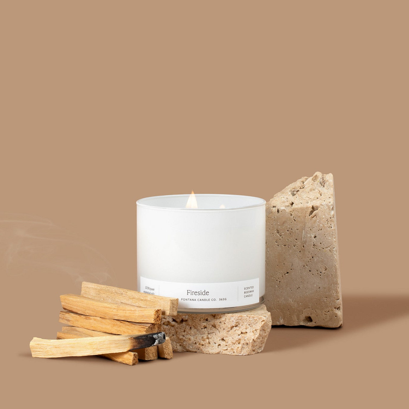 Lit 3-wick candle in a white glass vessel next to a pile of Palo Santo sticks in front of a tan background. 