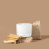 Lit 3-wick candle in a white glass vessel next to a pile of Palo Santo sticks in front of a tan background. 