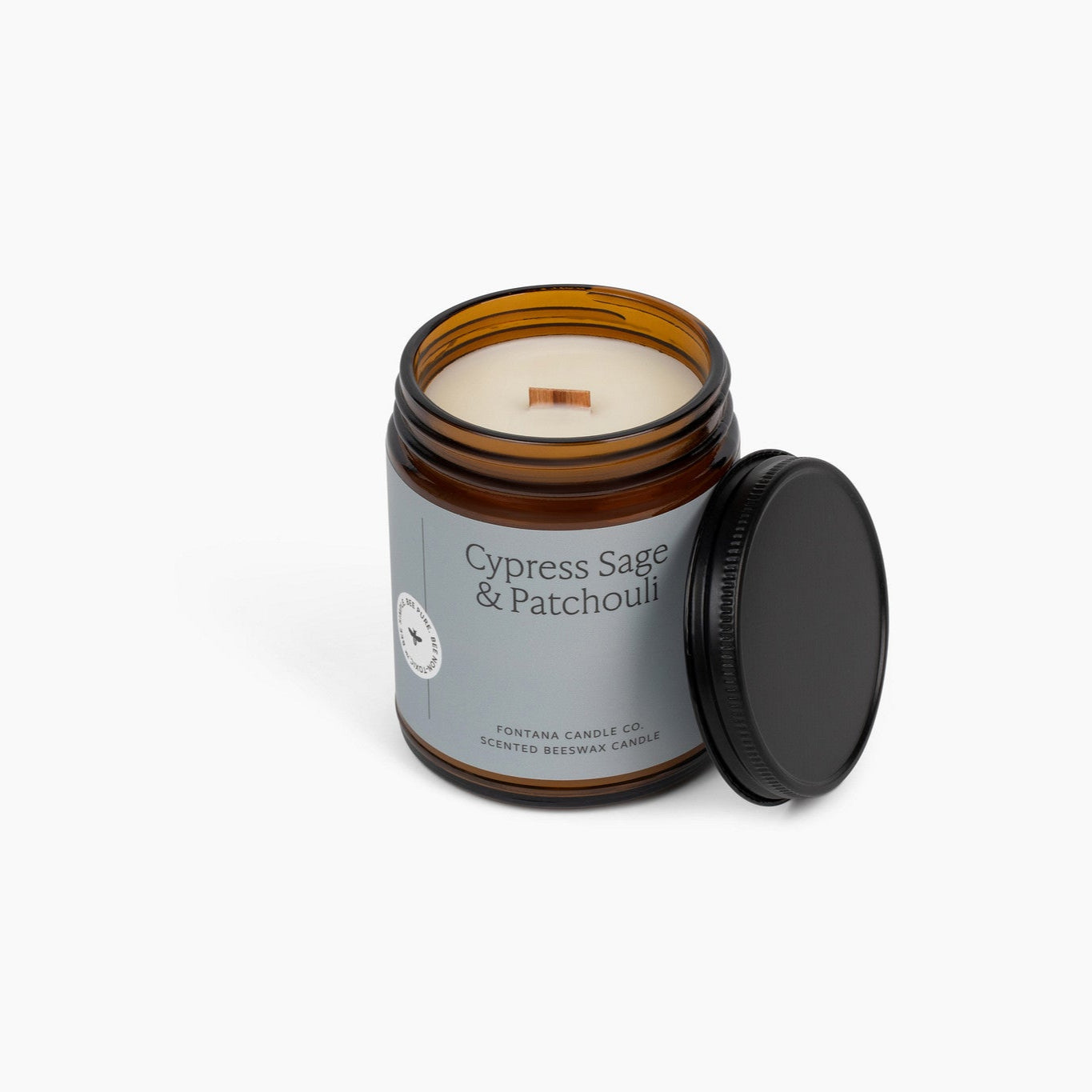 Unlit wooden wick candle, in an amber glass jar with a light blue label. This candle is in front of a white background. 