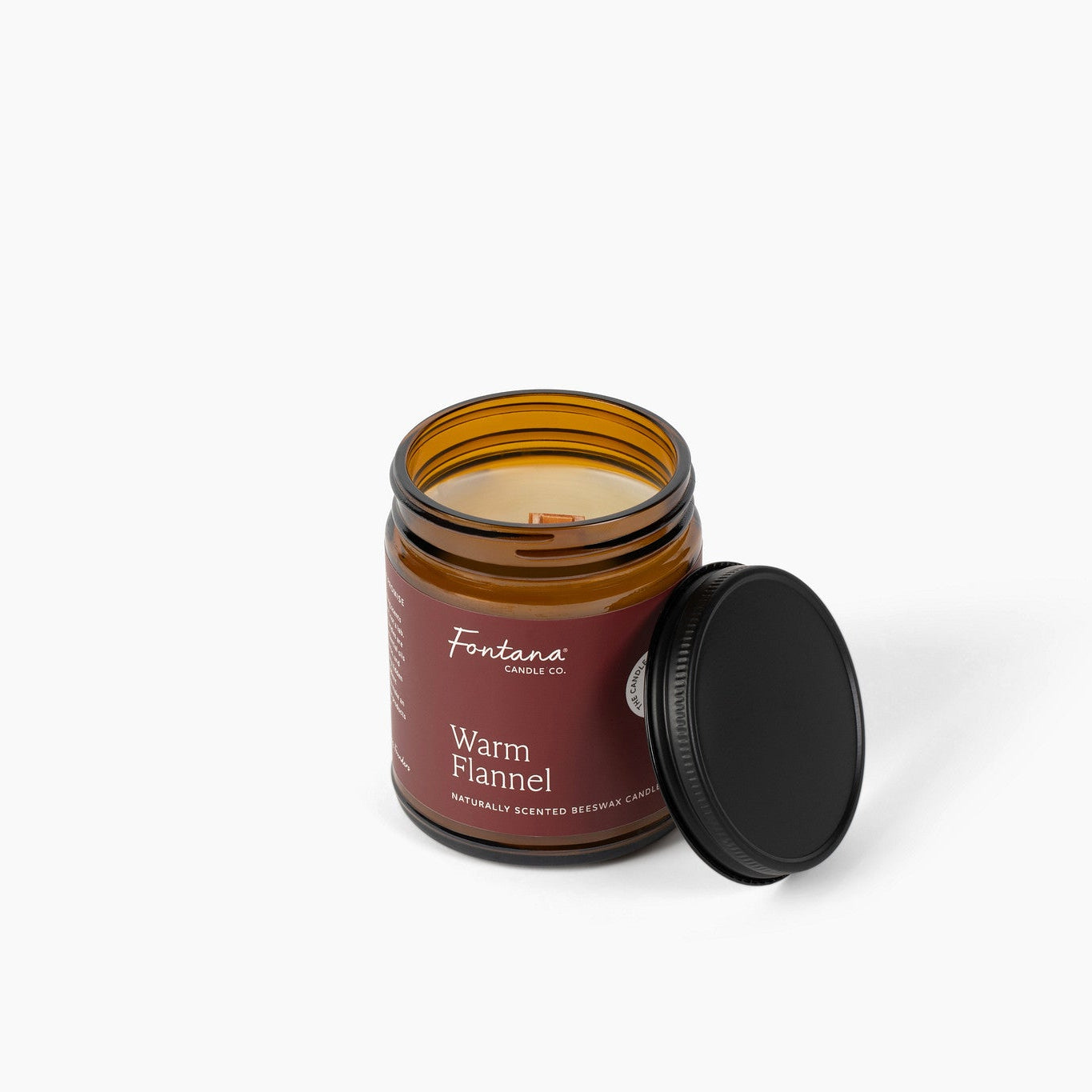 An amber glass jar candle with a black lid leaning against it on a clean white background. The label reads 'Fontana Candle Co' and 'Warm Flannel'.