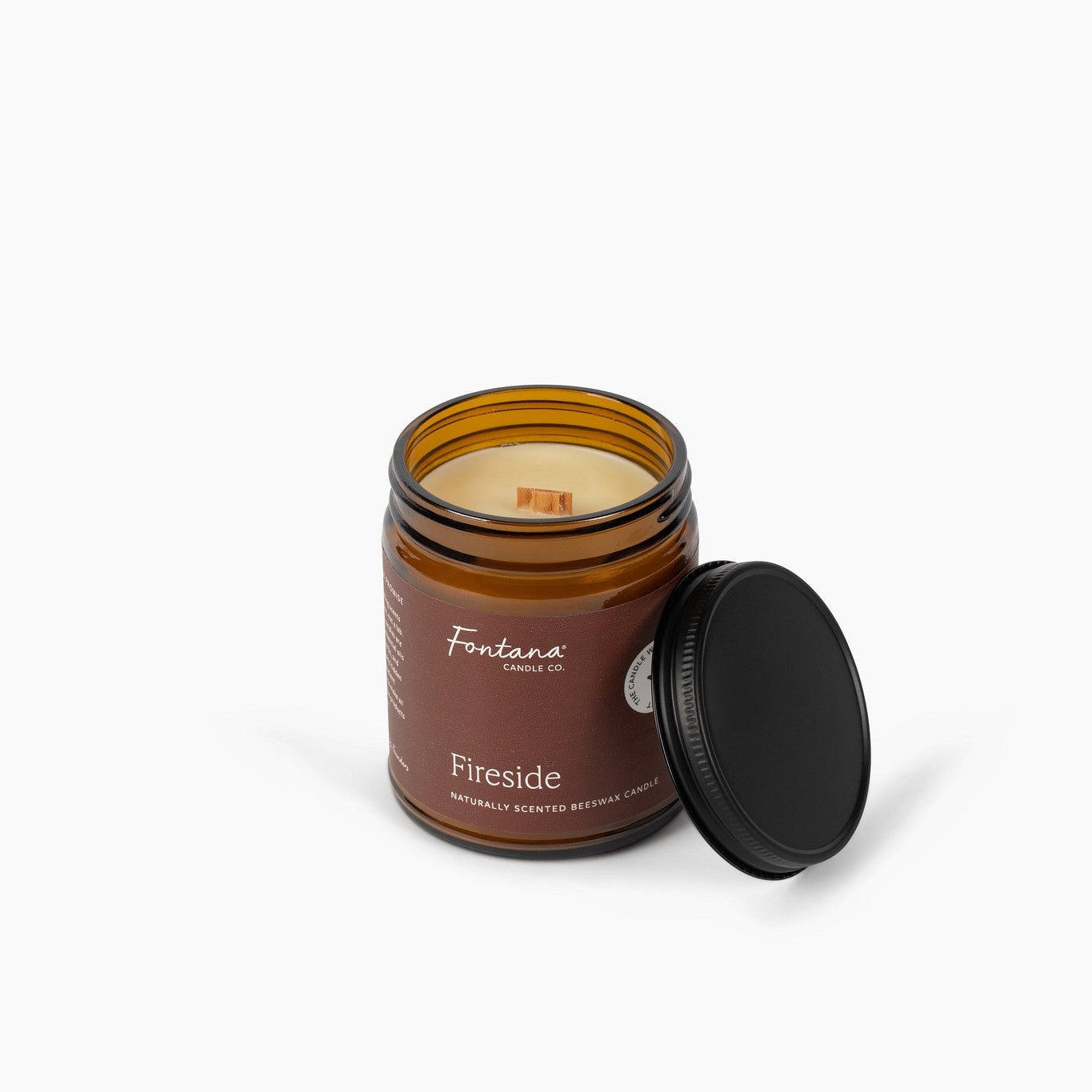 An amber glass jar candle with a black lid resting beside it on a clean white background. The label reads 'Fontana Candle Co' and 'Fireside'.