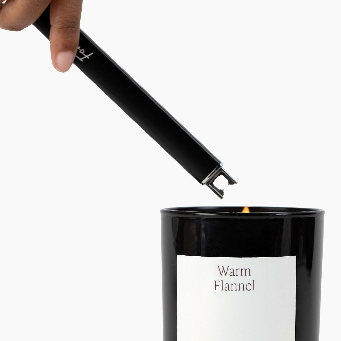 Hand using a black candle lighter to light a candle labeled 'Warm Flannel' on a white background