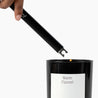 Hand using a black candle lighter to light a candle labeled 'Warm Flannel' on a white background