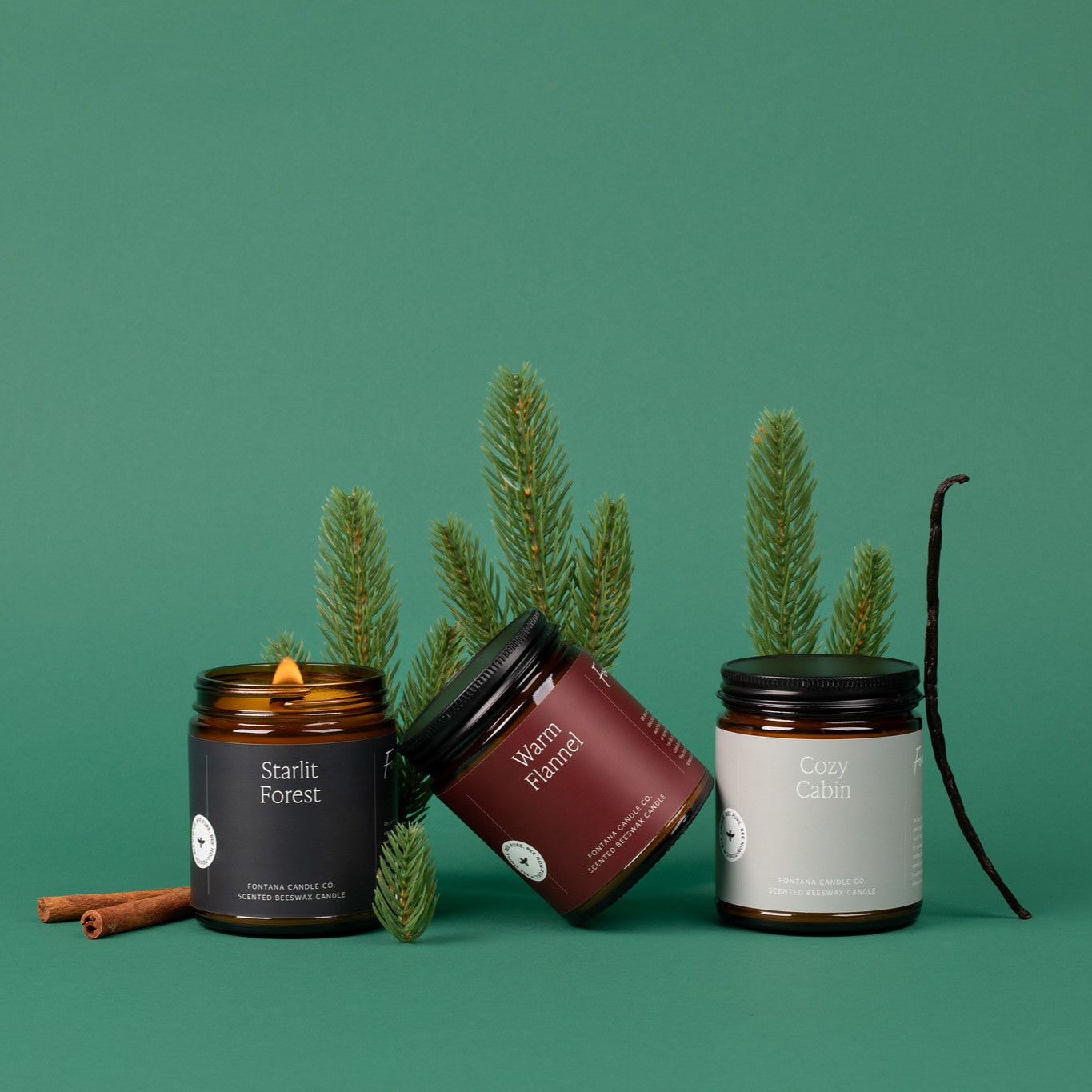 Three amber glass jar candles on a green background surrounded by fir needles, cinnamon sticks, and vanilla beans. 
