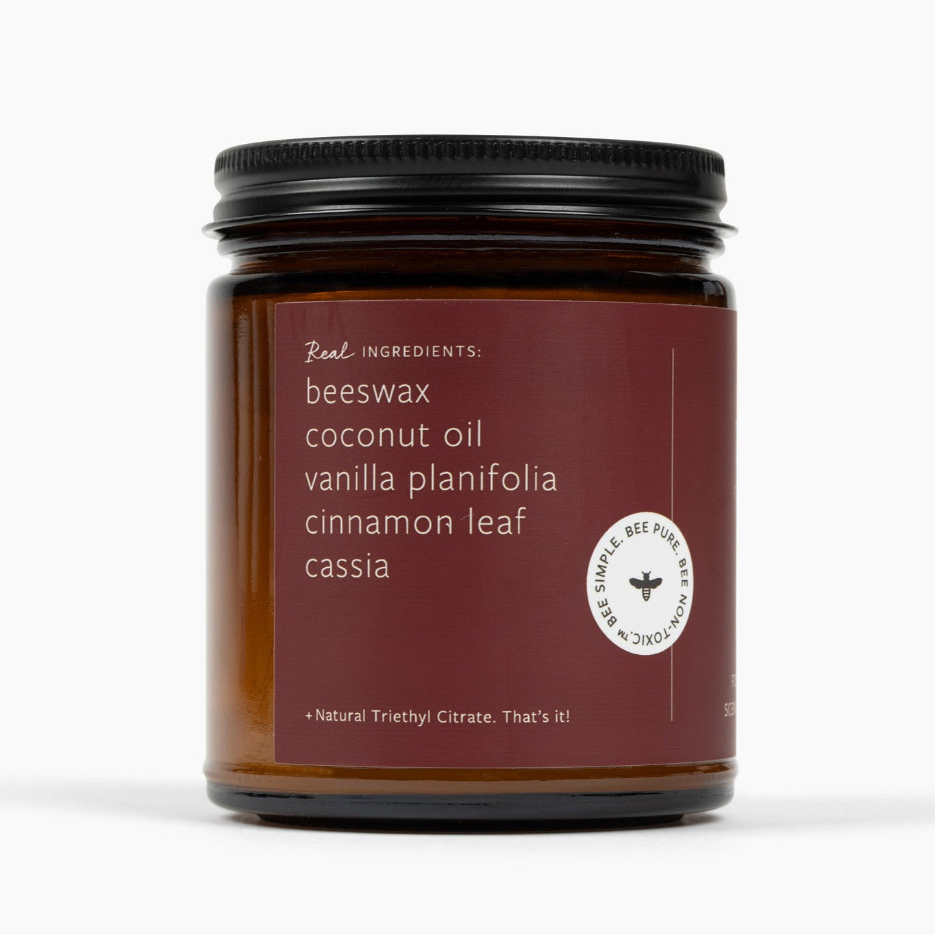 A side shot of an amber glass jar candle with a deep red label and the ingredients: beeswax, coconut oil, vanilla planifolia, cinnamon leaf, cassia, and natural triethyl citrate.
