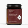 A side shot of an amber glass jar candle with a deep red label and the ingredients: beeswax, coconut oil, vanilla planifolia, cinnamon leaf, cassia, and natural triethyl citrate.