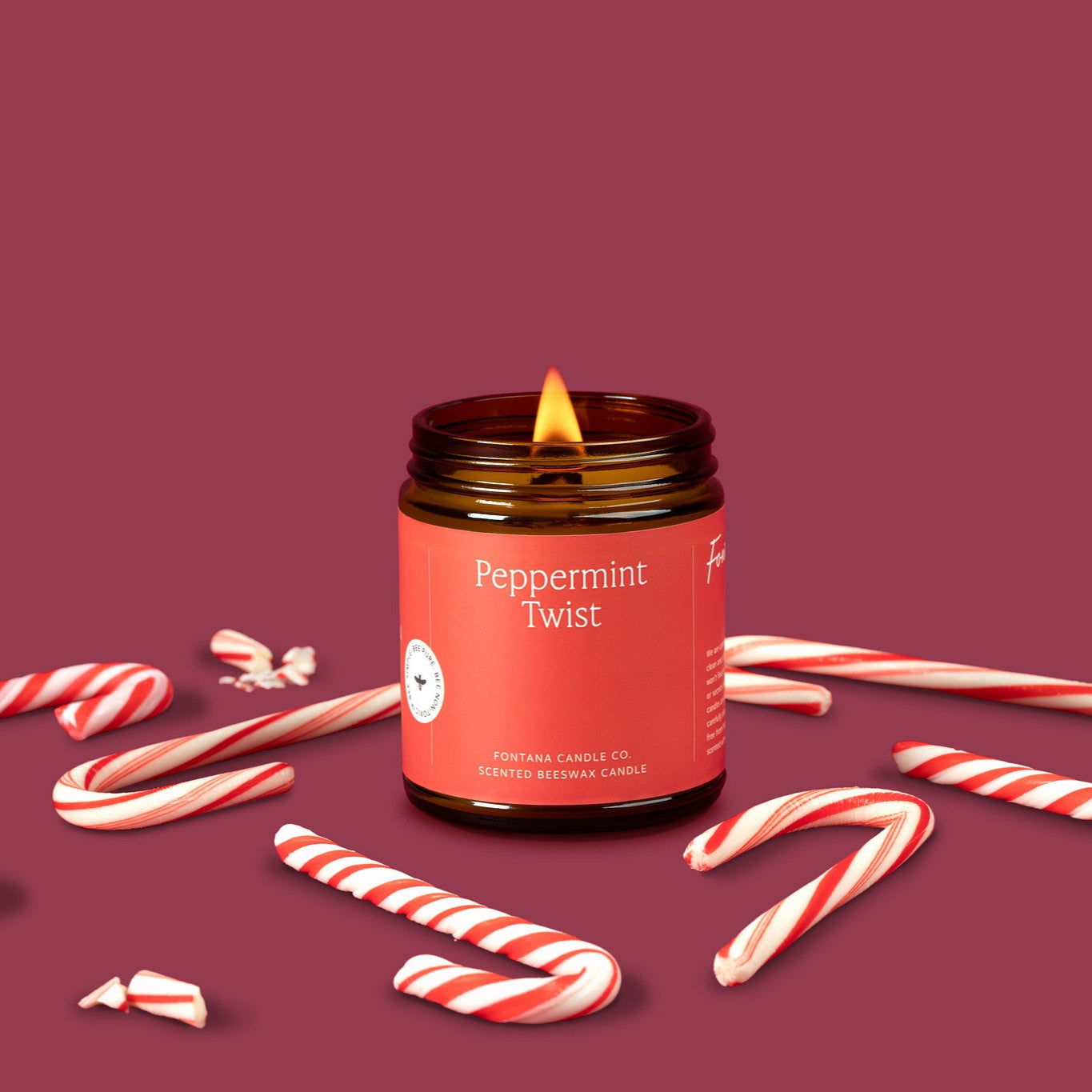 Amber glass jar candle labeled 'Peppermint Twist' surrounded by candy canes on a maroon background.