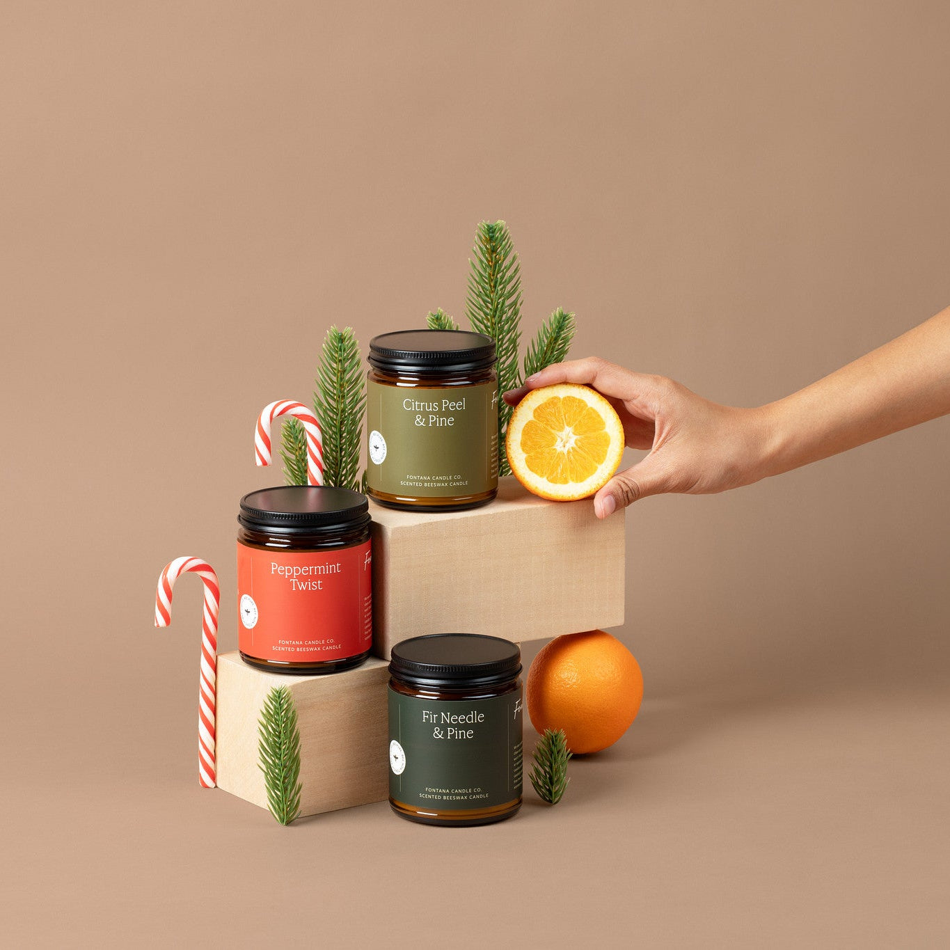 Three amber glass jar candles with a red, light green, and dark green label, surrounded by candy canes, evergreen branches, and oranges. 