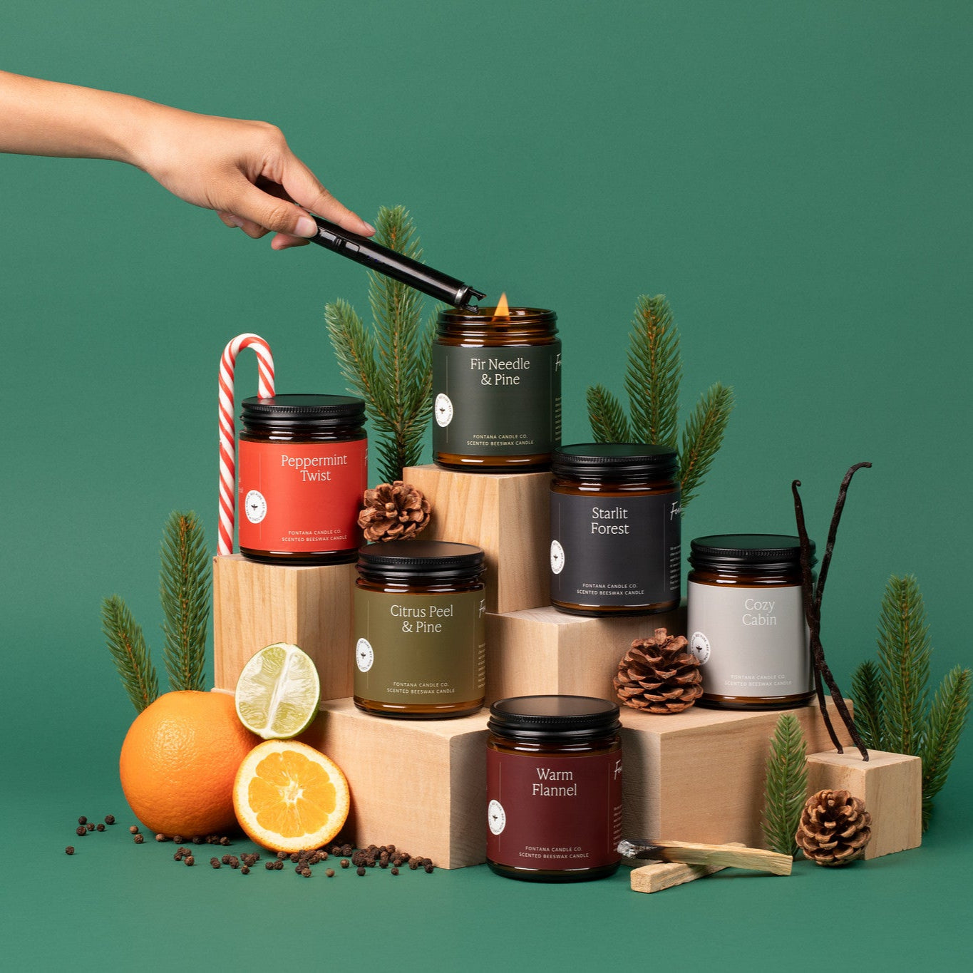 6 amber glass jar holiday candles sitting on wooden blocks, surrounded by oranges, spices, palo santo, candy canes, pinecones, and pine branches, in front of a green background.