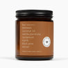 Side of an amber glass jar candle with a deep brown label and the ingredients: beeswax, coconut oil, vanilla planifolia, cardamom, myrrh, black pine, vetiver, and natural triethyl citrate.