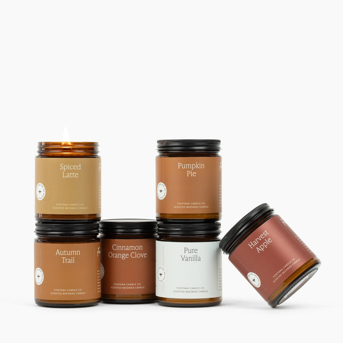 Six amber glass jar candles on a white background: Spiced Latte, Autumn Trail, Cinnamon Orange Clove, Pumpkin Pie, Pure Vanilla, Harvest Apple.