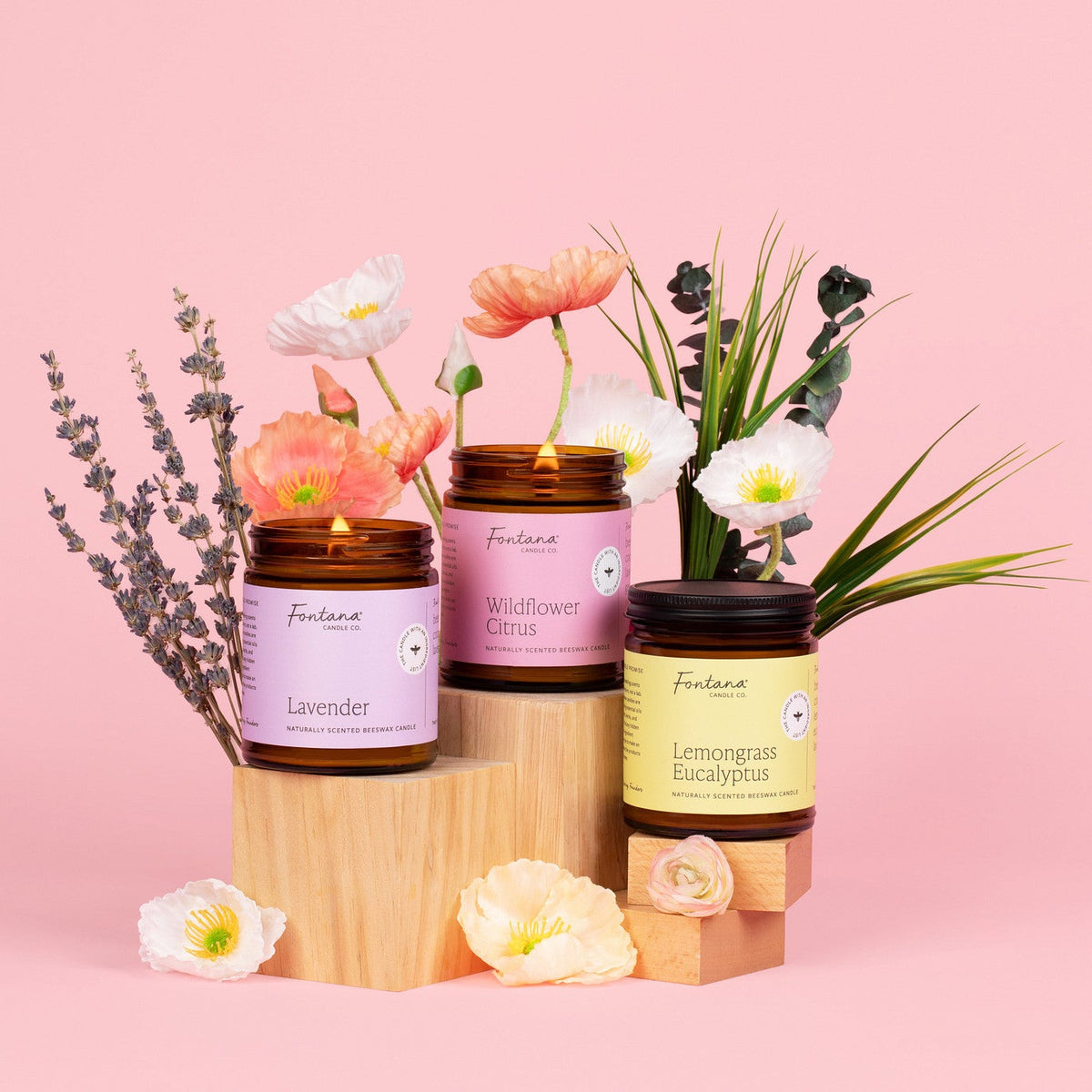 three 9oz jar candles, lavender, wildflower citrus, lemongrass eucalyptus, posed around a variety of flowers and lavender on a pink background