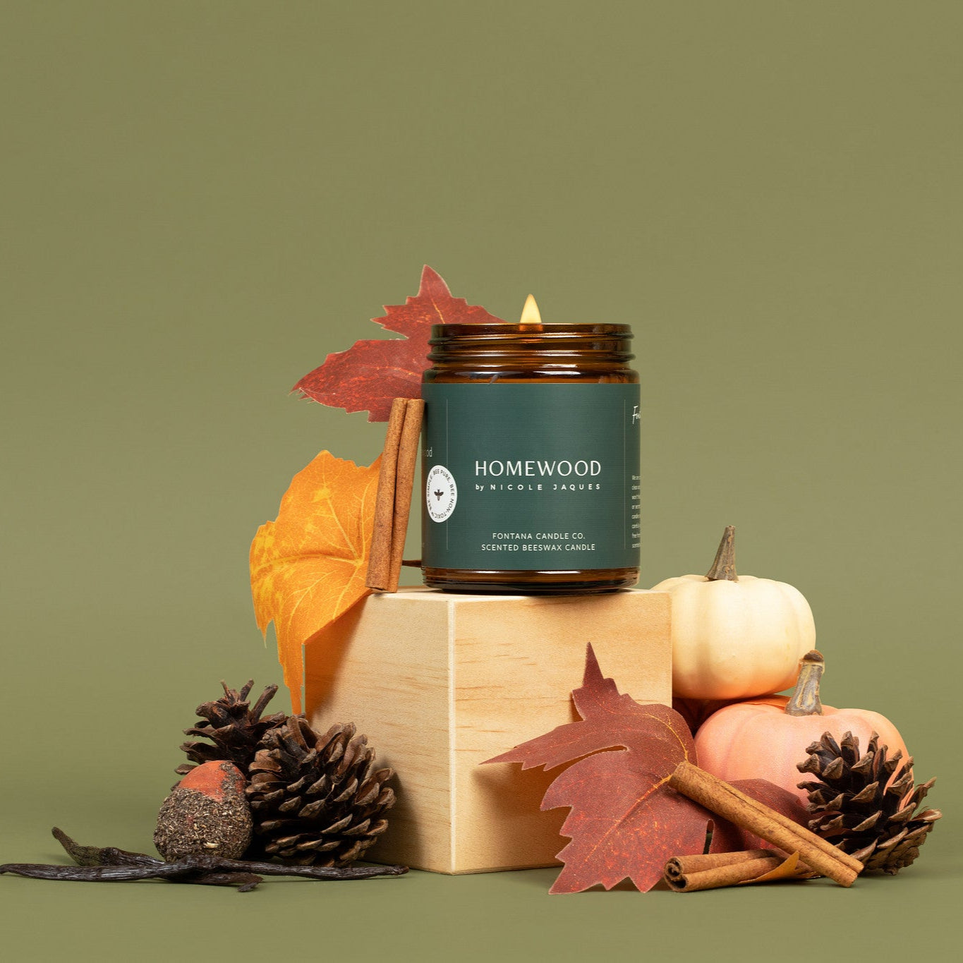 Homewood amber glass jar candle with autumn-themed decor on a green background.