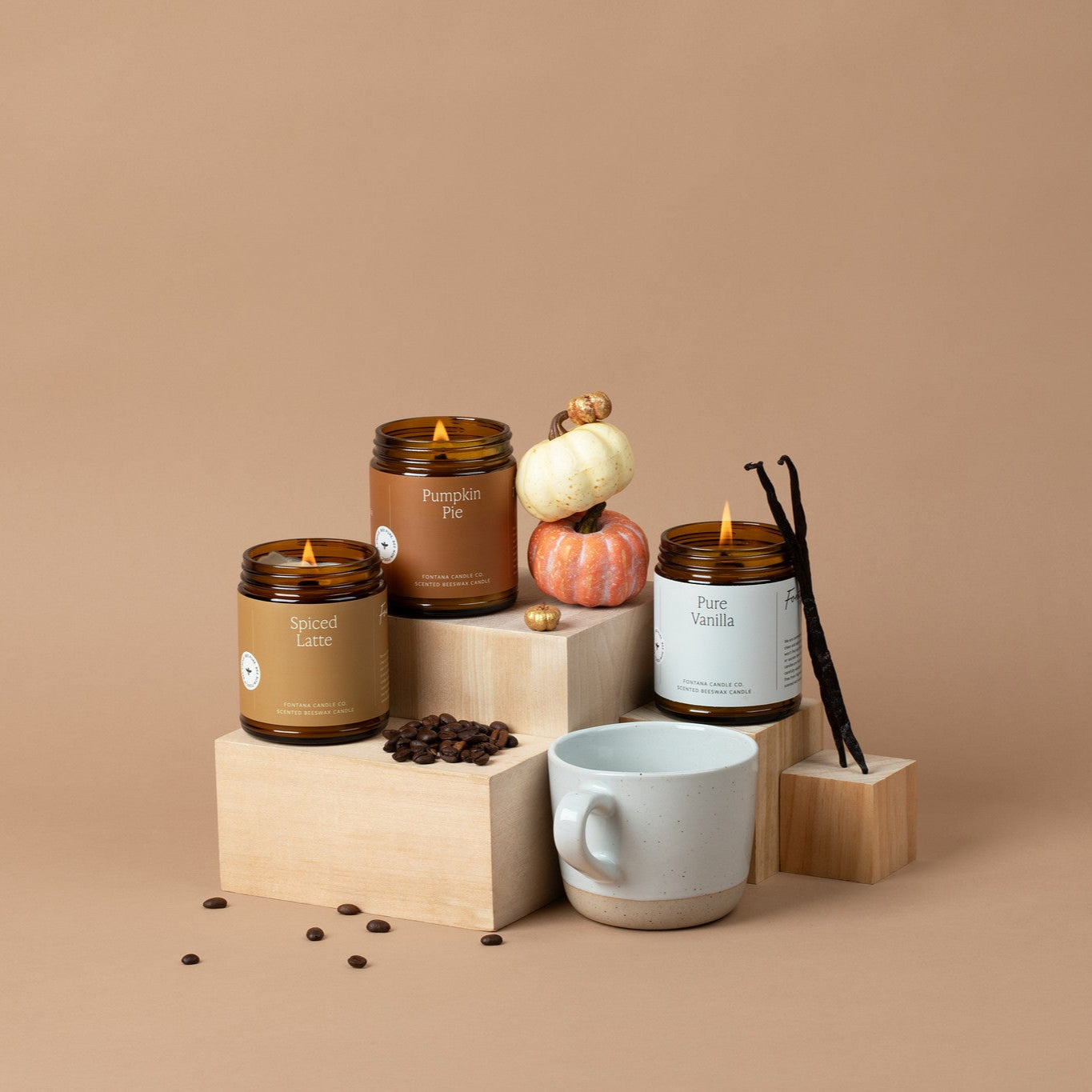 3 lit amber glass jar candles in front of a tan background, surrounded by coffee beans, a coffee mug, pumpkins, and vanilla beans.