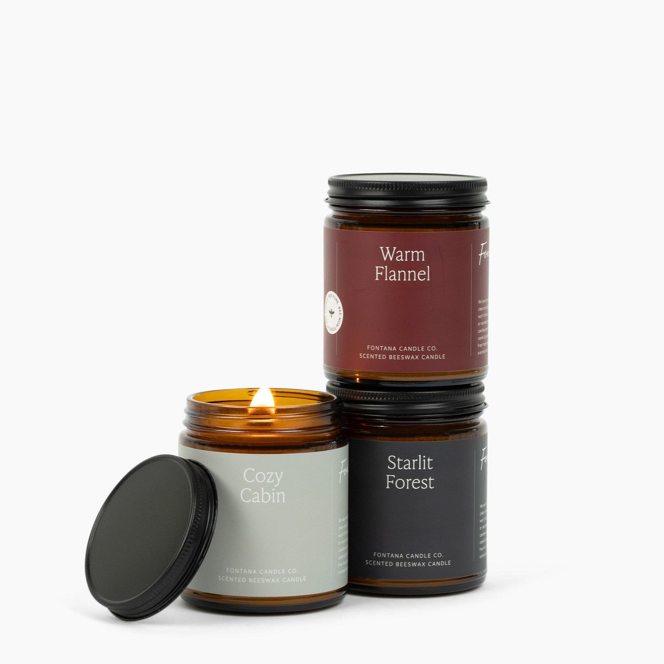 Three amber glass jar candles on a white background with the scent names: Cozy Cabin, Starlit Forest, and Warm Flannel. 