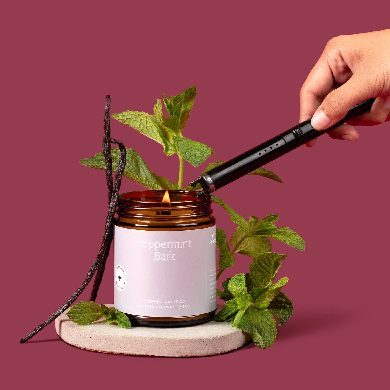Lit amber glass jar with a gray label in front of a deep purple background and surrounded by mint leaves and vanilla beans. 