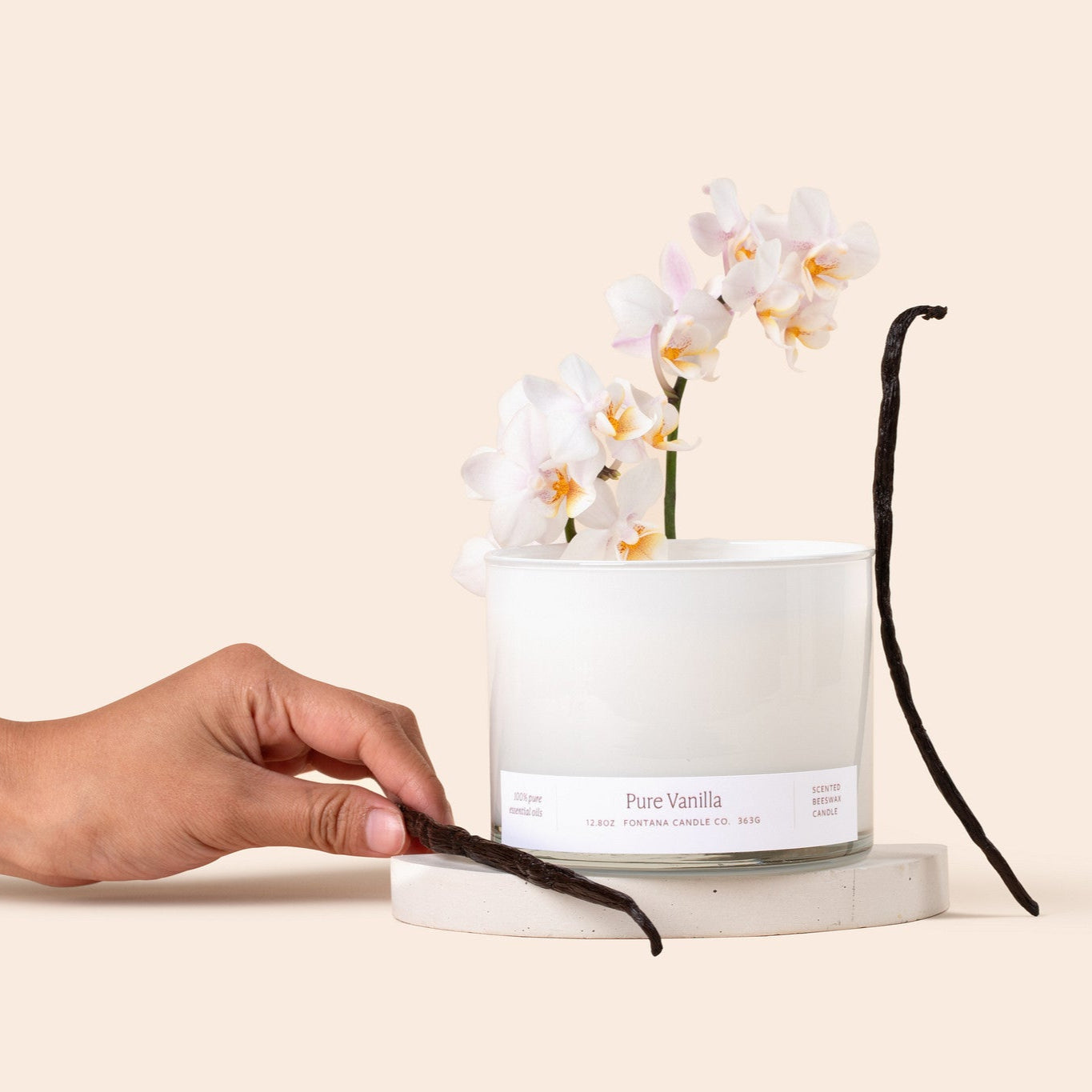 3-wick candle in a white glass vessel in front of a beige background and surrounded by white orchids and vanilla pads. 