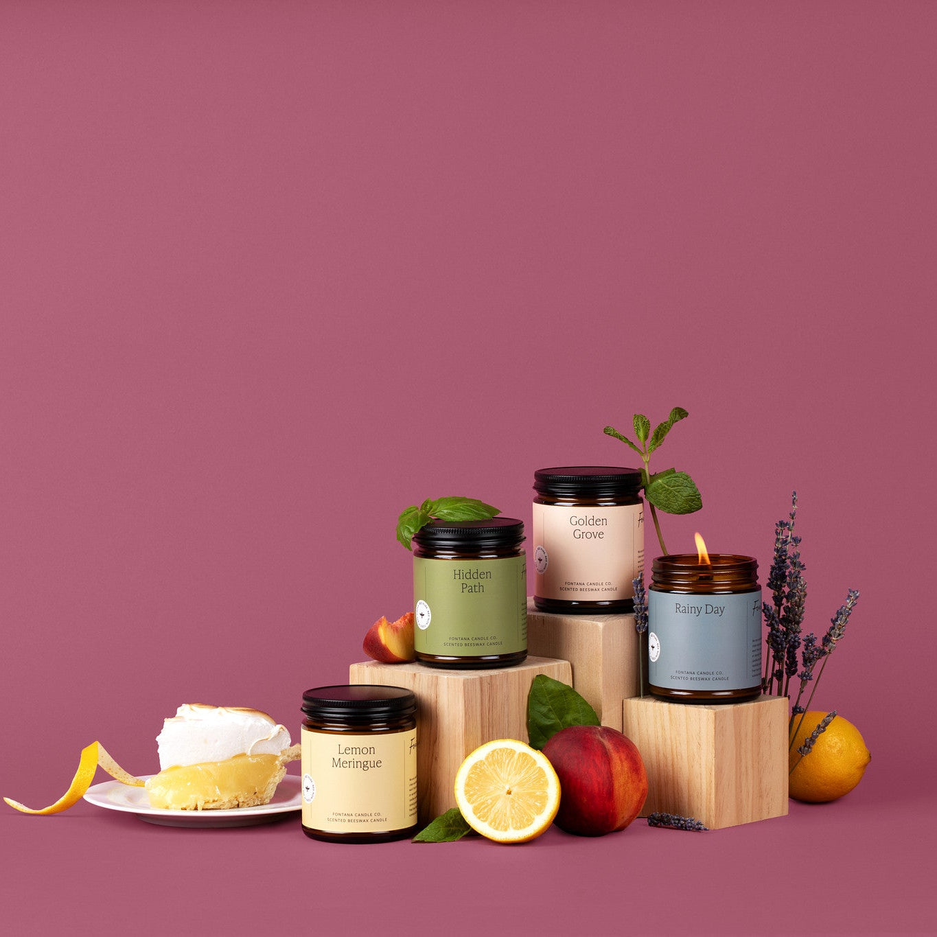 4 amber glass jar candles with colorful labels in front of a burgendy background with flowers, citrus fruit, and a lemon meringue pie. 