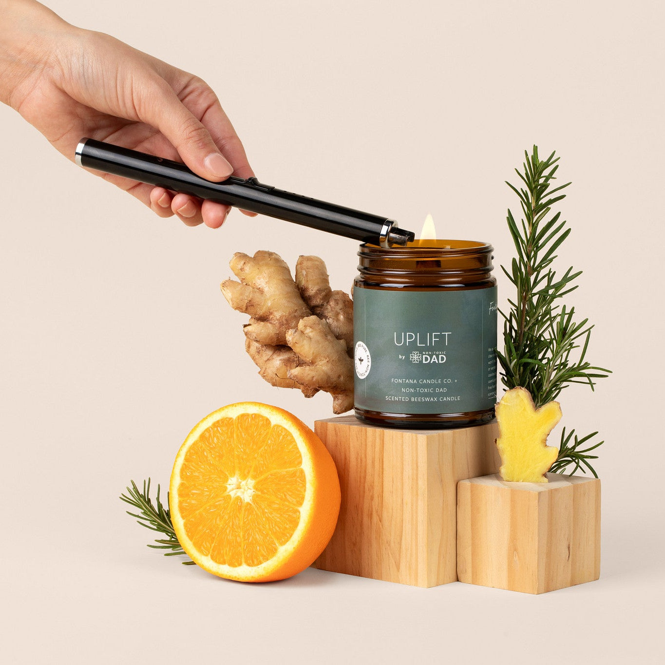 Hand lighting an amber glassjar candle with ginger, orange, and rosemary on a beige background