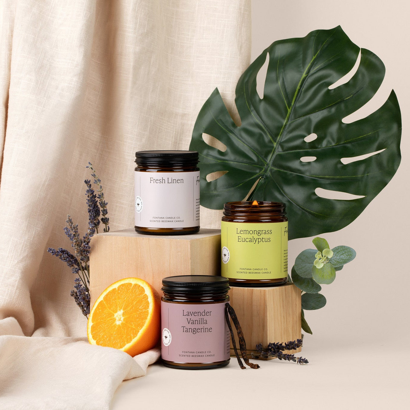 Three amber glass jar candles with colorful labels on a beige background, surrounded by lavender, mint, vanilla pods, oranges, and a big green leaf.